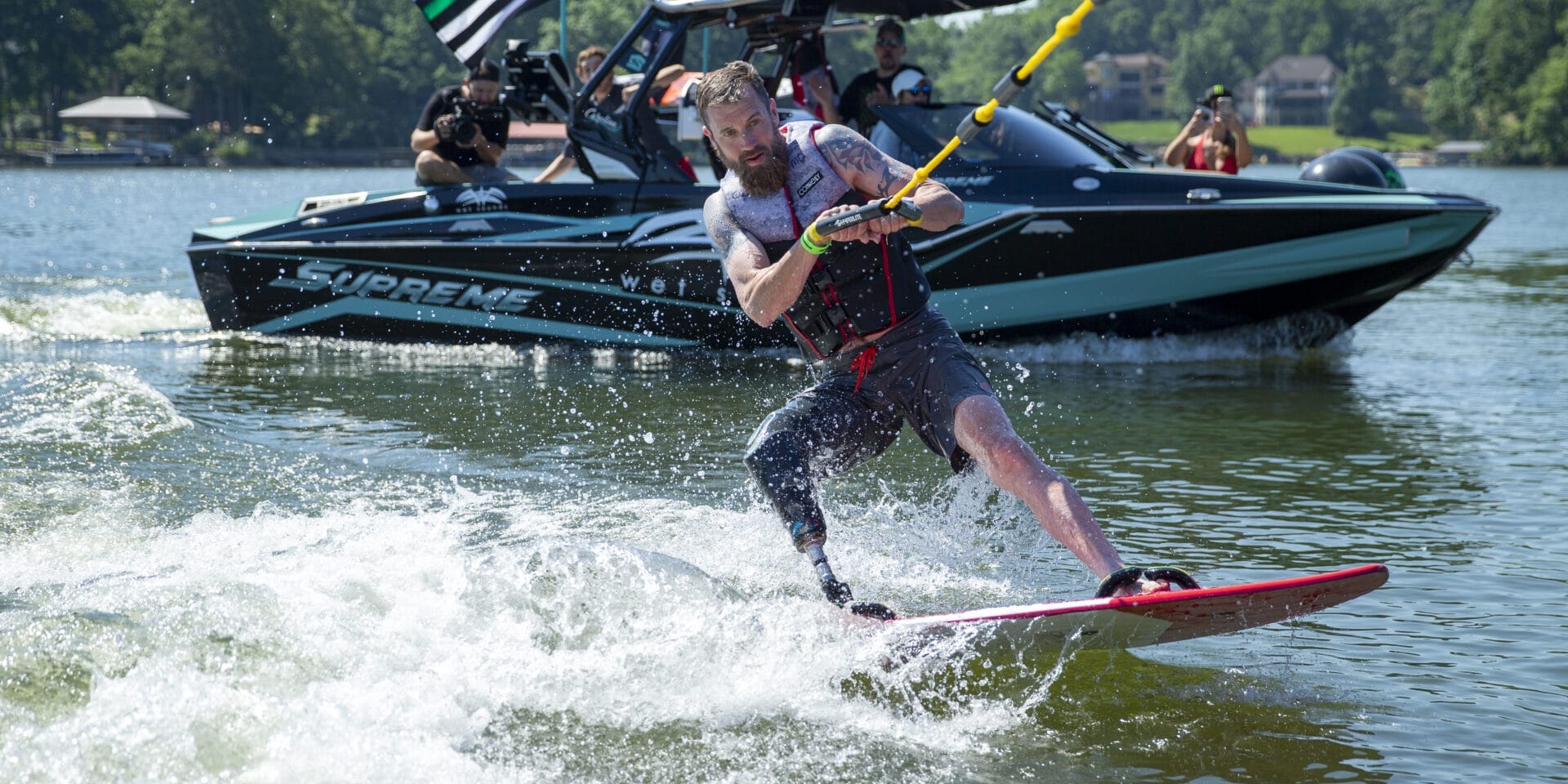 A man wakeboards on a lake, holding a rope, with a boat carrying people and flags in the background.