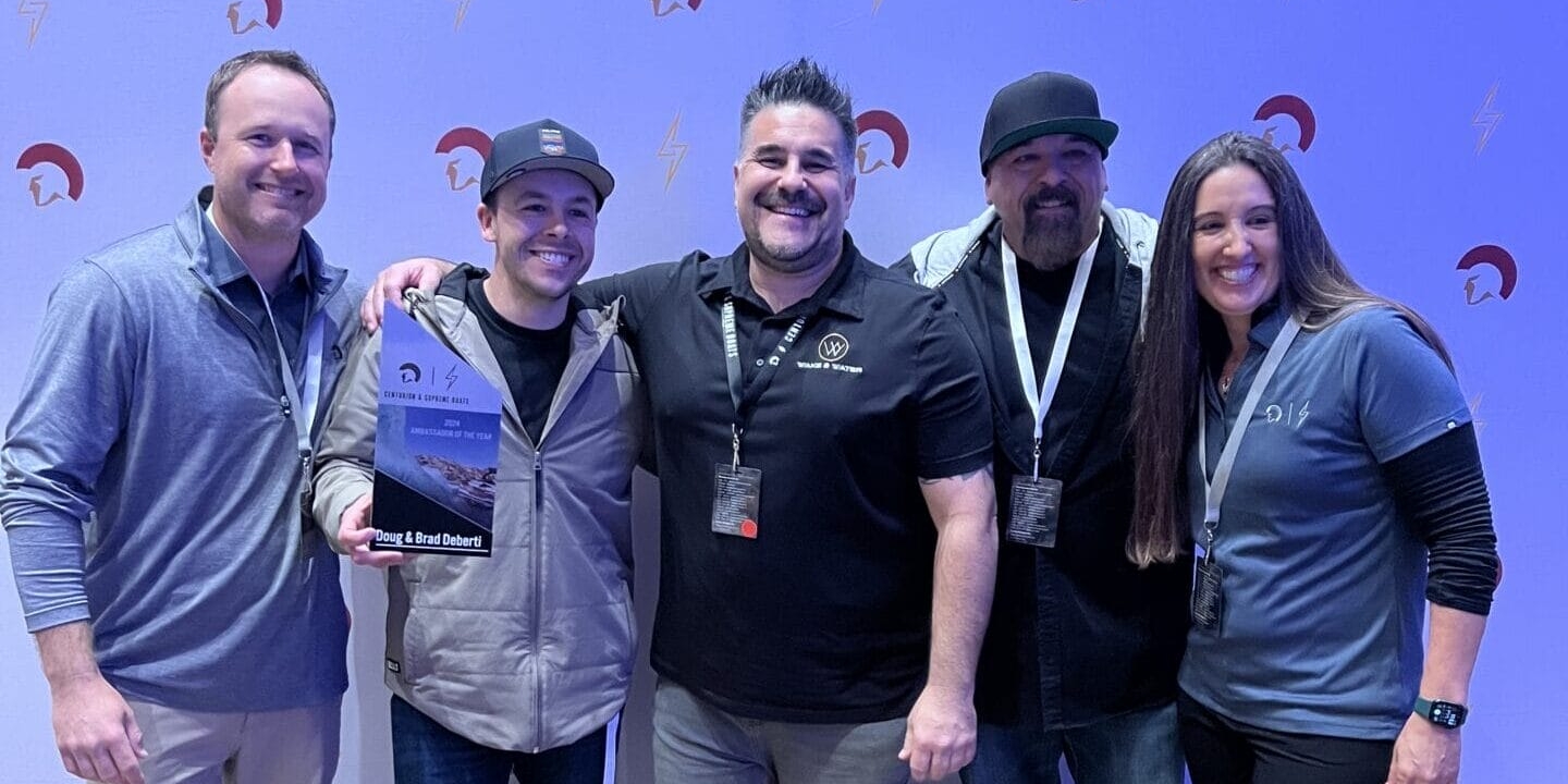 Five people standing together smiling, one person holding a trophy. They are in a room with a patterned carpet and a backdrop featuring logos.