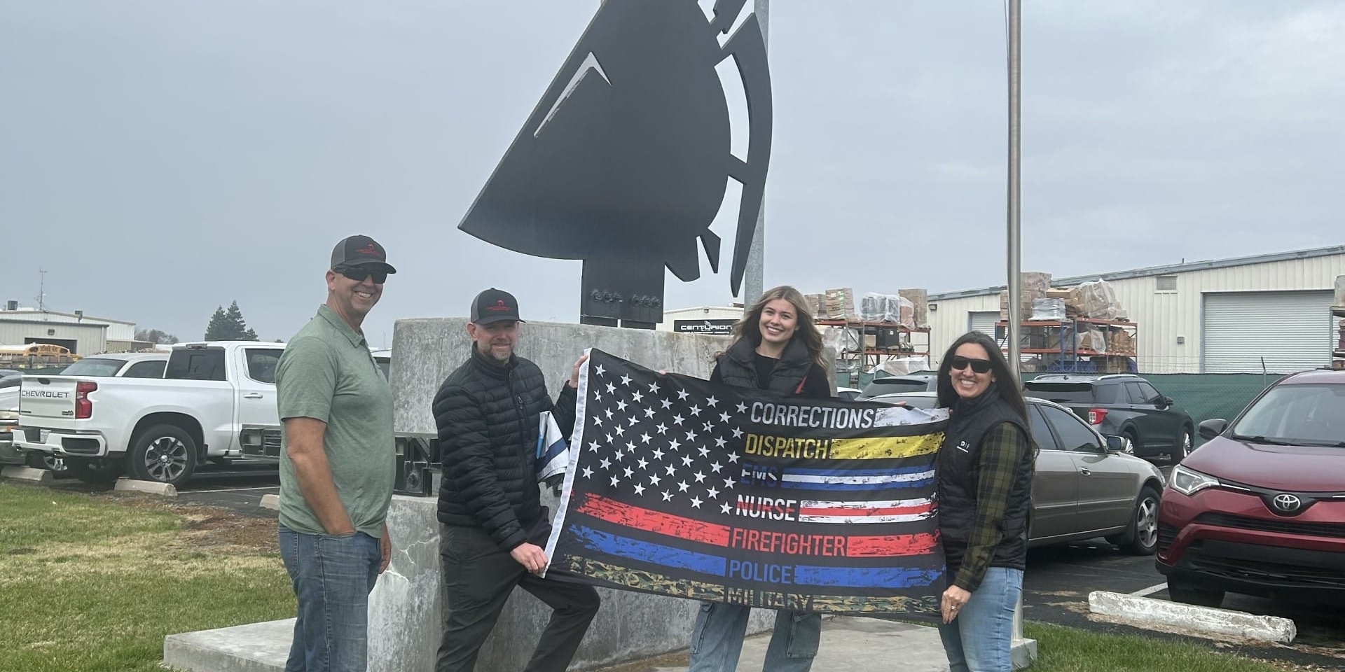 Four individuals hold a flag with various service stripes in front of a Spartan helmet statue. Vehicles and a flagpole are in the background.