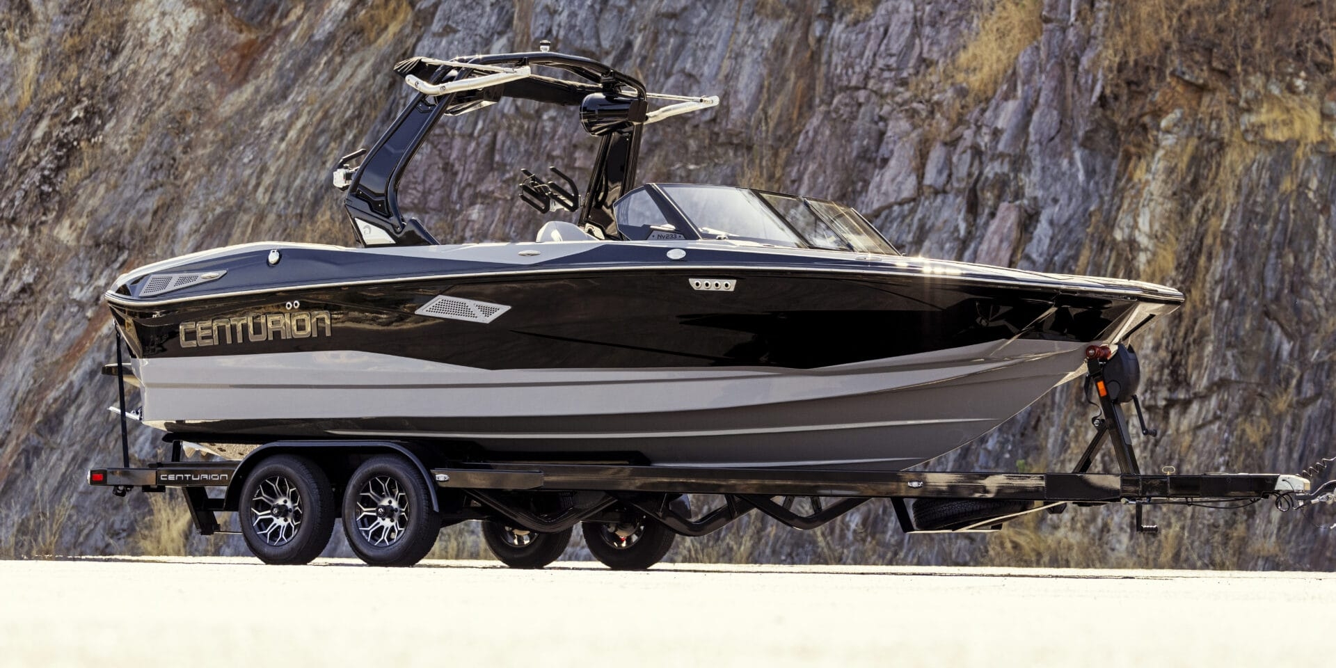 Side view of a black and white Centurion boat on a trailer, parked on a road with a rocky cliff background.