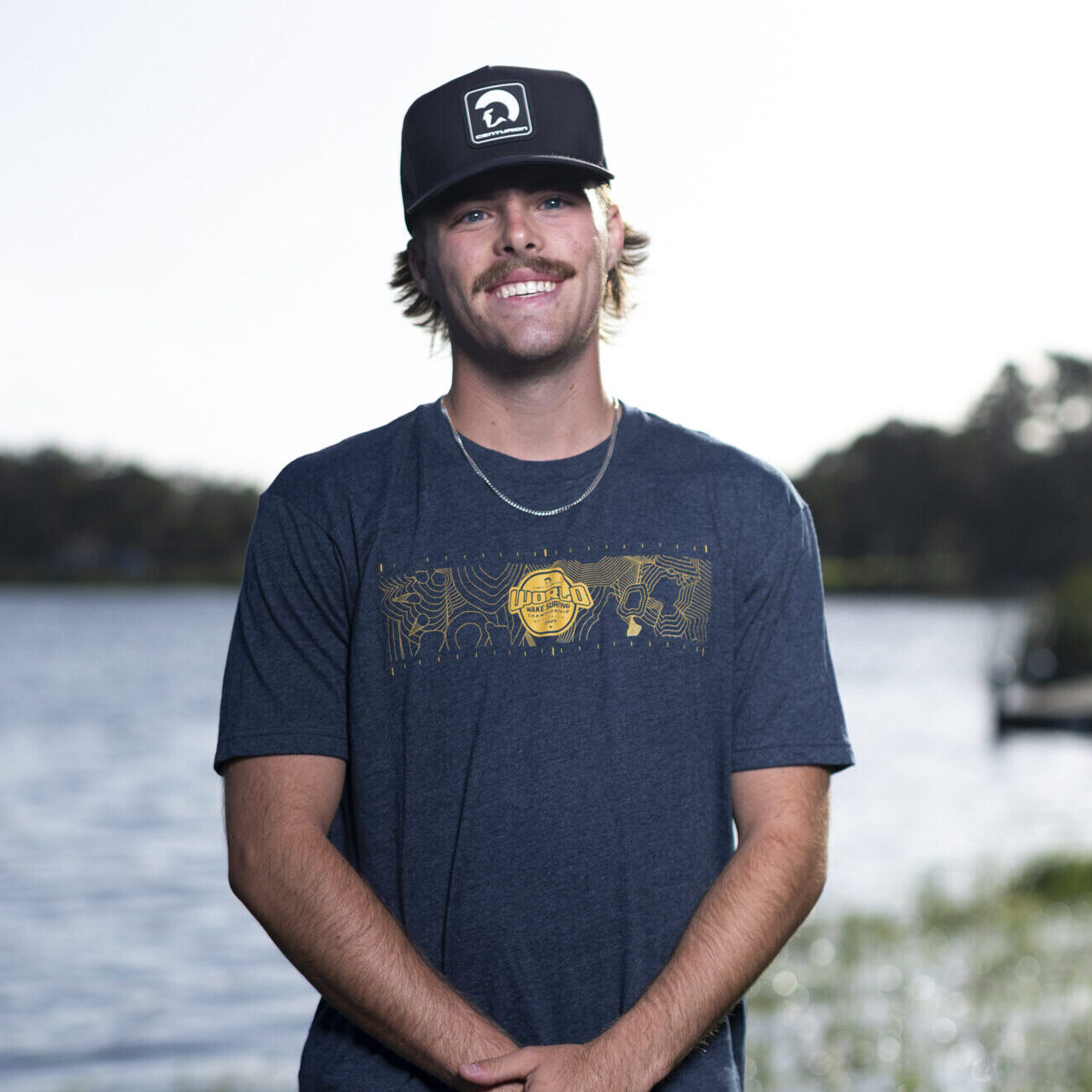 A young man with a mustache wearing a dark baseball cap, navy T-shirt, and patterned shorts stands near a lake with greenery in the background.