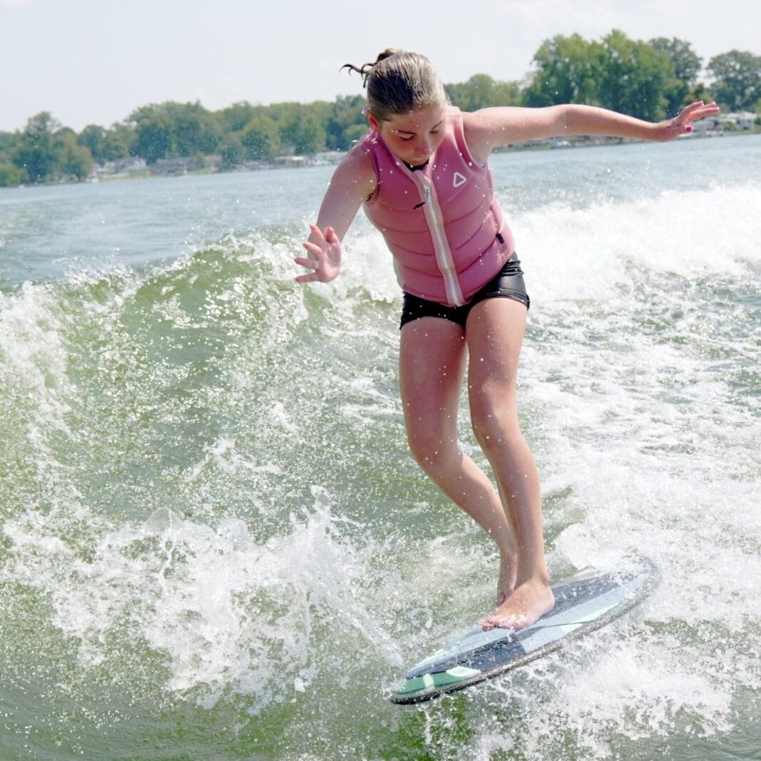 Person surfing on a wave in a lake, wearing a pink life jacket and black shorts, with trees visible in the background.