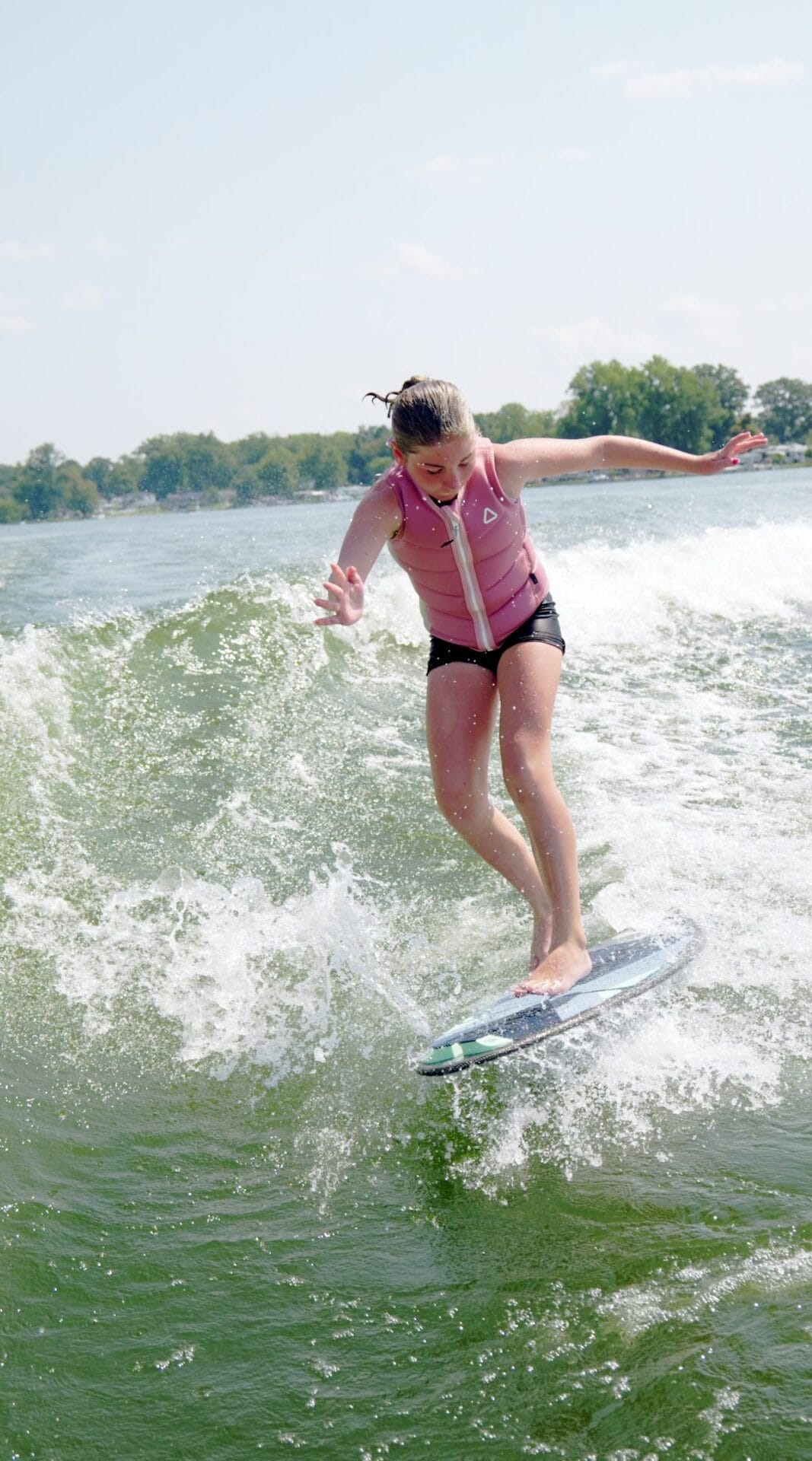 Person surfing on a wave in a lake, wearing a pink life jacket and black shorts, with trees visible in the background.