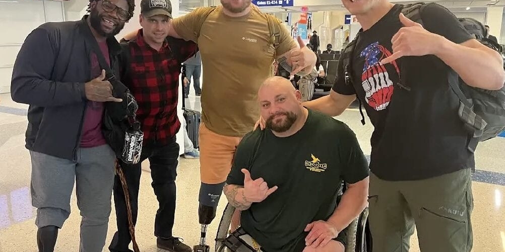 A group of five men, some with prosthetic legs, pose and smile at an airport terminal. They are casually dressed and making hand gestures. Airport signage is visible in the background.
