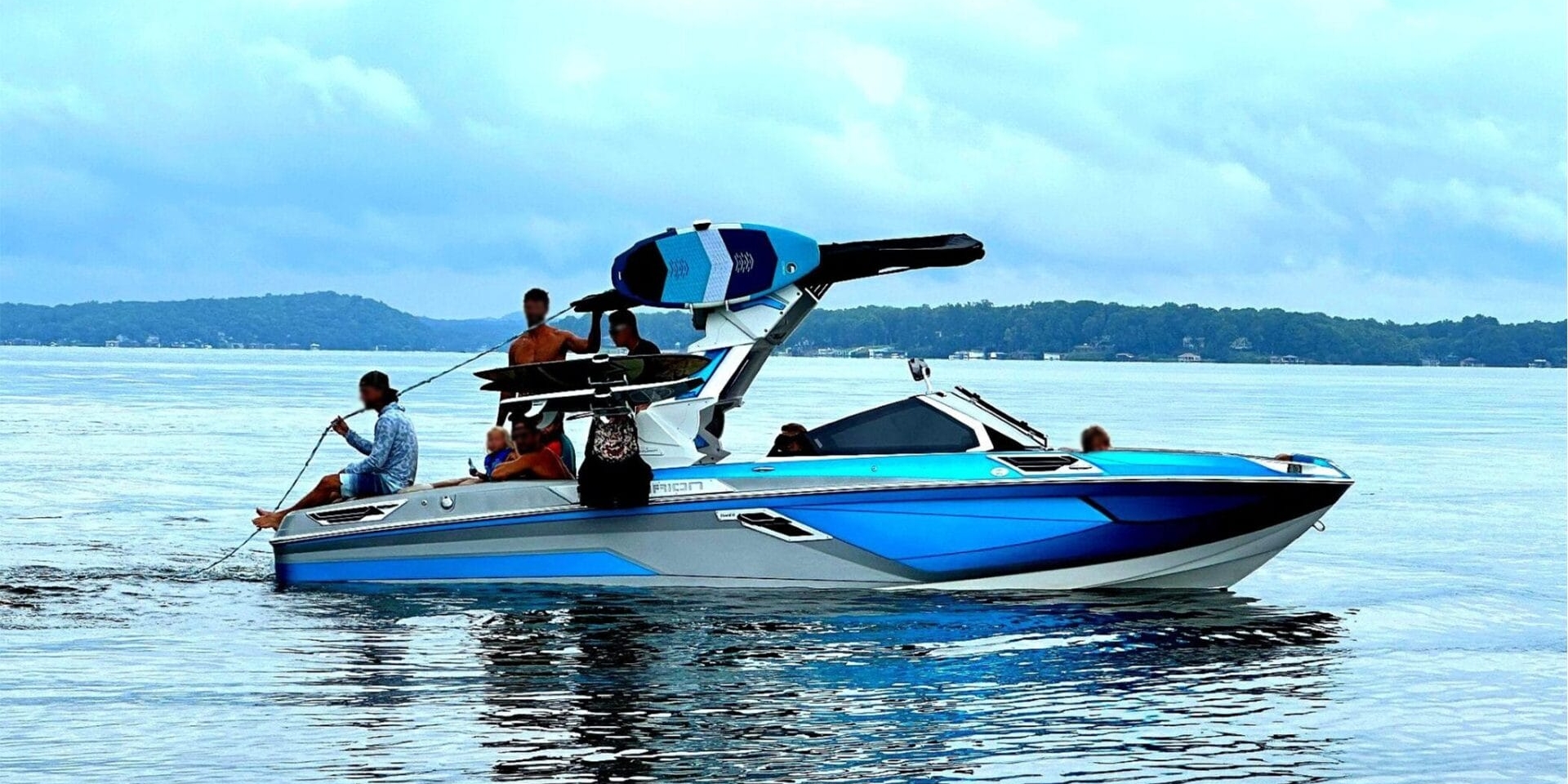 A blue and white speedboat with multiple people, including one holding a rope, is on a lake. Wakeboards are visible on the boat's rack. Trees and hills can be seen in the background under a cloudy sky.