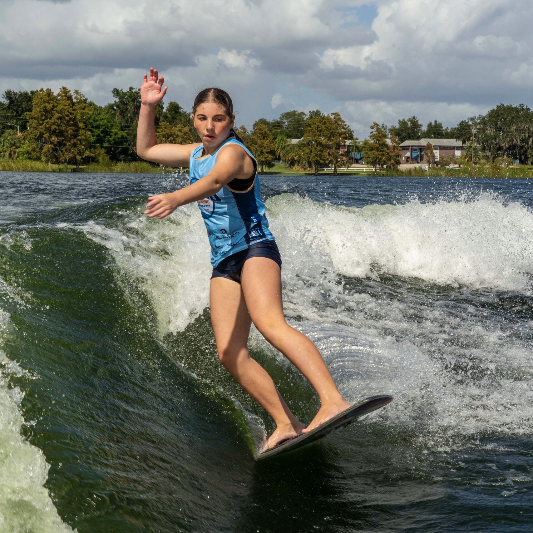 Aria Hellemn, dressed in blue, rides a wave on a surfboard under a partly cloudy sky, with trees and buildings in the background.