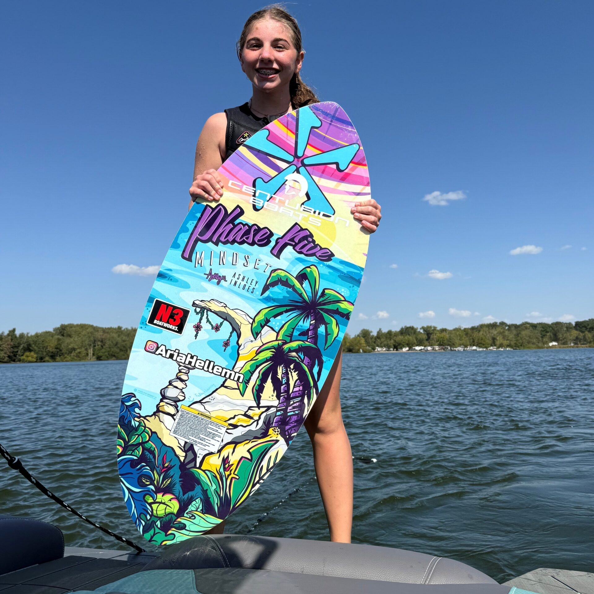 Teen girl Aria Hellemn stands on a boat, holding a colorful wakesurf board, with a lake and trees in the background under a clear blue sky.