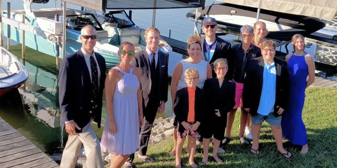 A group of people dressed in formal and casual attire pose by a dock with boats in the background.