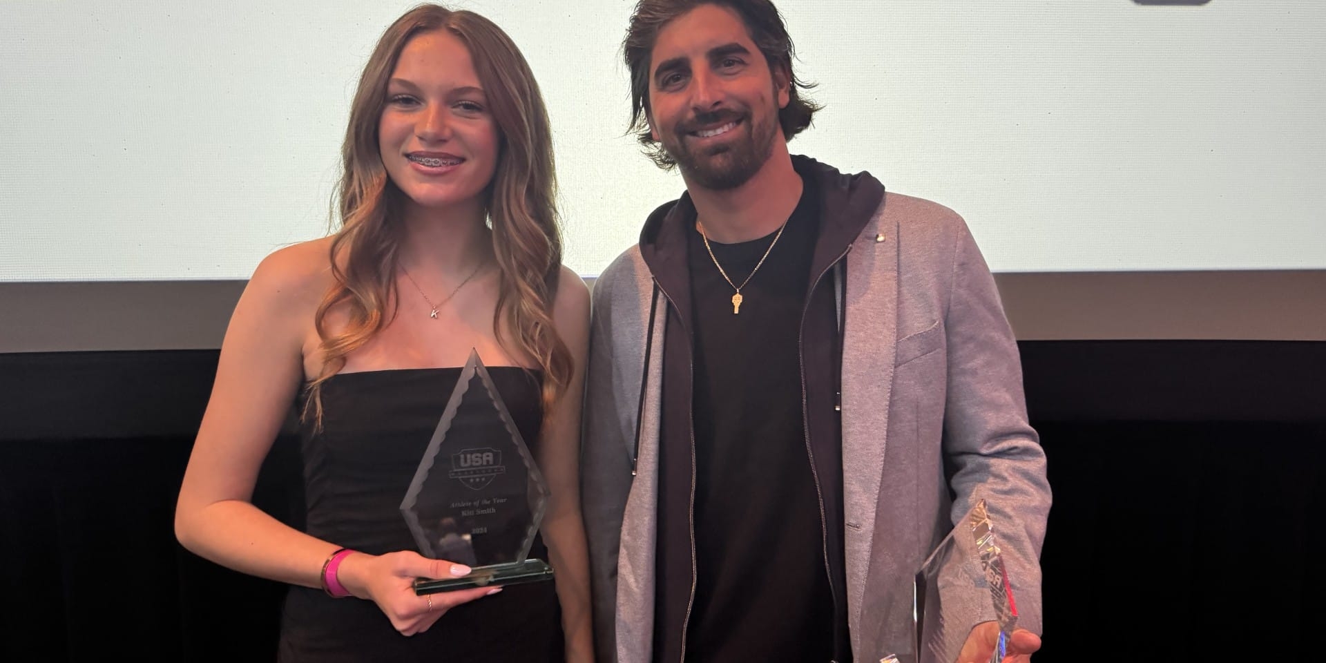Two people holding awards and smiling at a banquet, standing in front of a backdrop that reads 