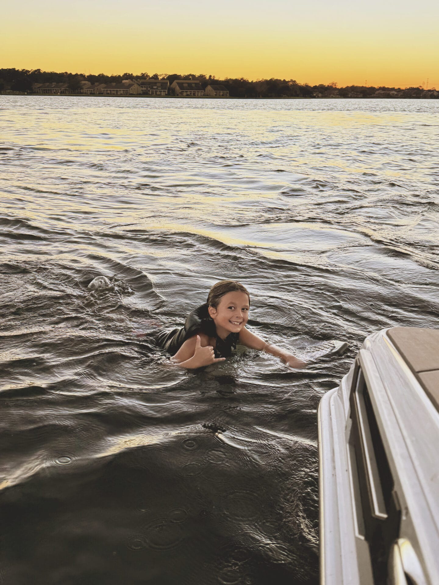 Child swimming in a lake at sunset, wearing a life vest and making a shaka sign with one hand near a boat's edge.