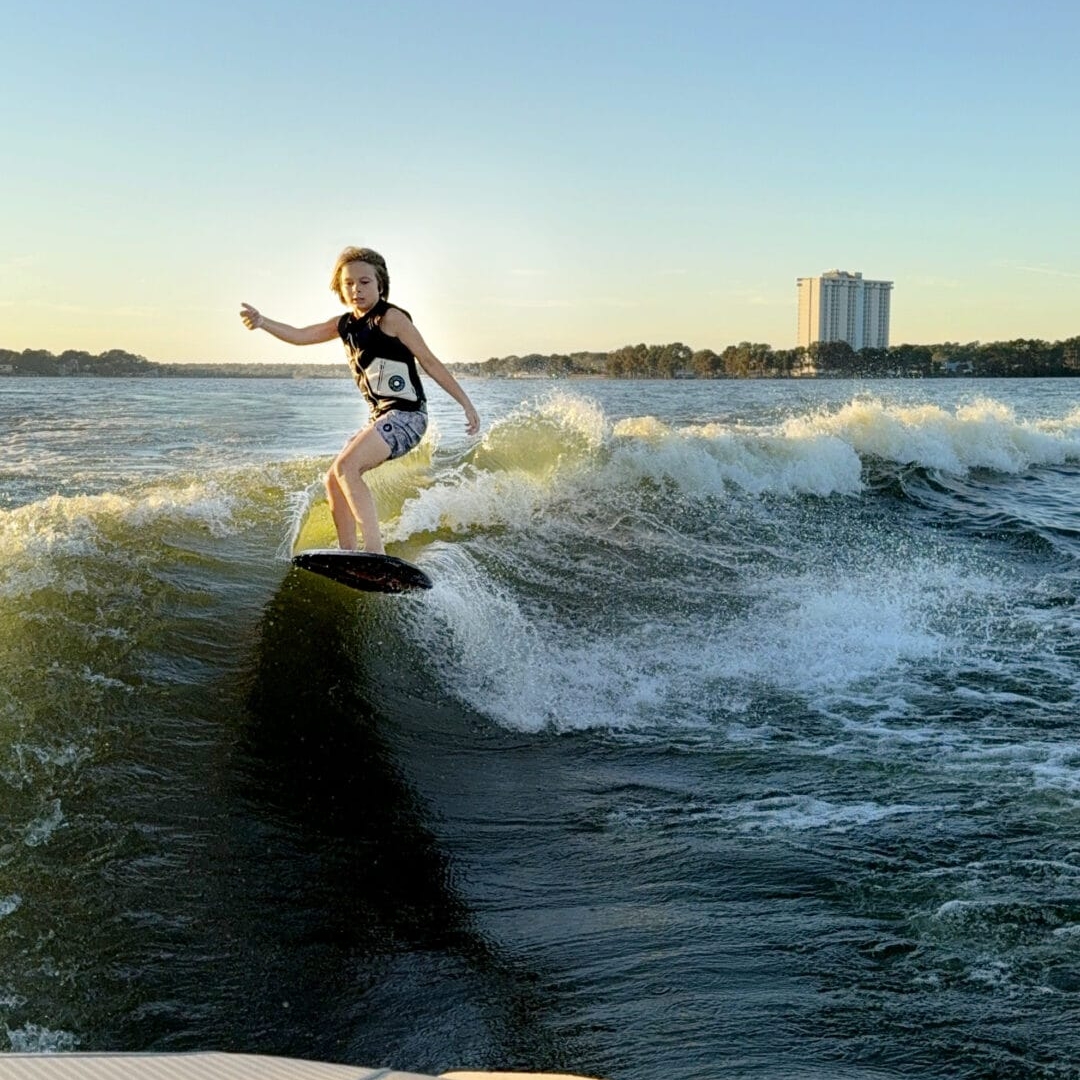 Person wearing a helmet rides a surfboard on a wave behind a boat at sunset, with a large building visible in the background.