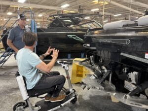 Two men converse near black boats inside a warehouse. One man, seated on a mobility scooter, gestures with his hands.