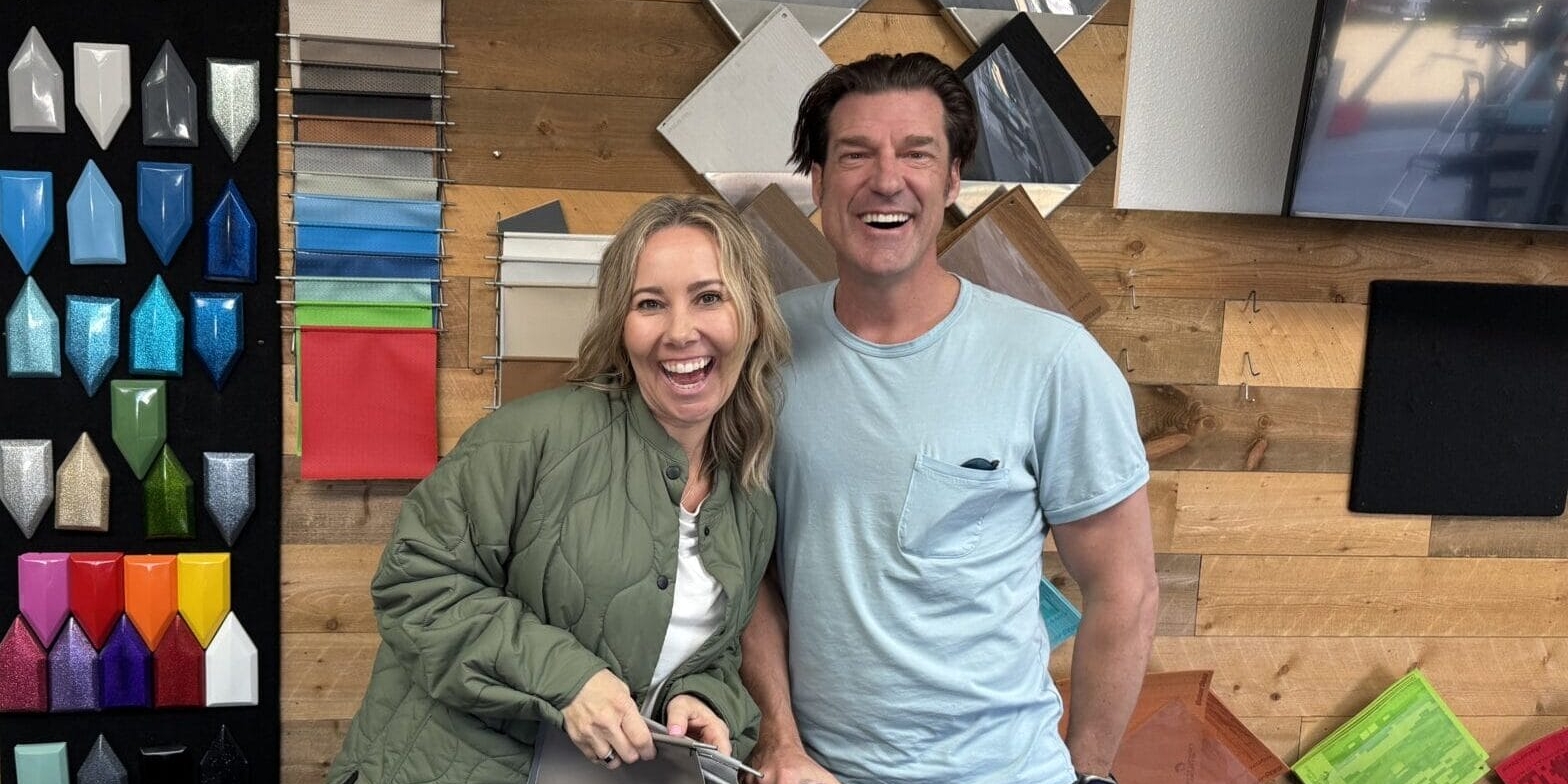 Two people smiling inside a room with samples of colorful materials displayed on the wall behind them.