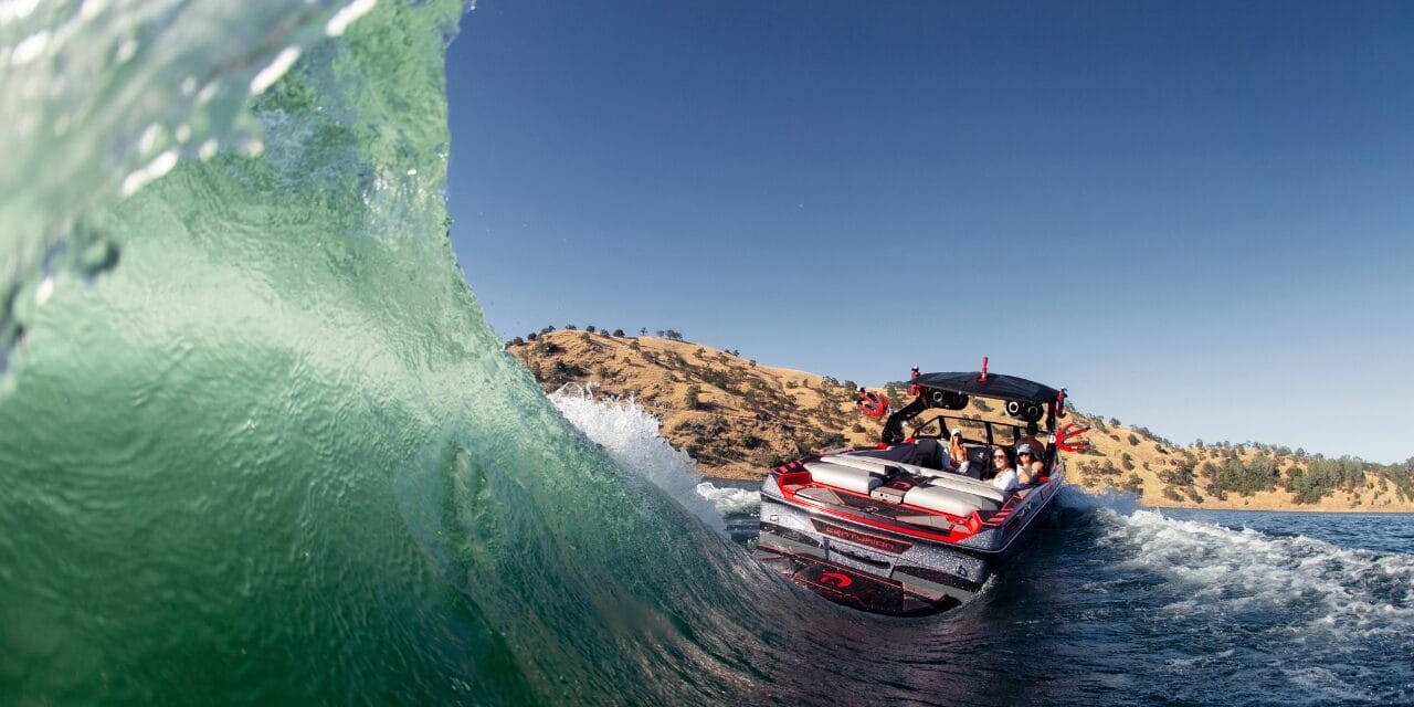 A speedboat with passengers navigates through a wave on a sunny day with hills in the background.