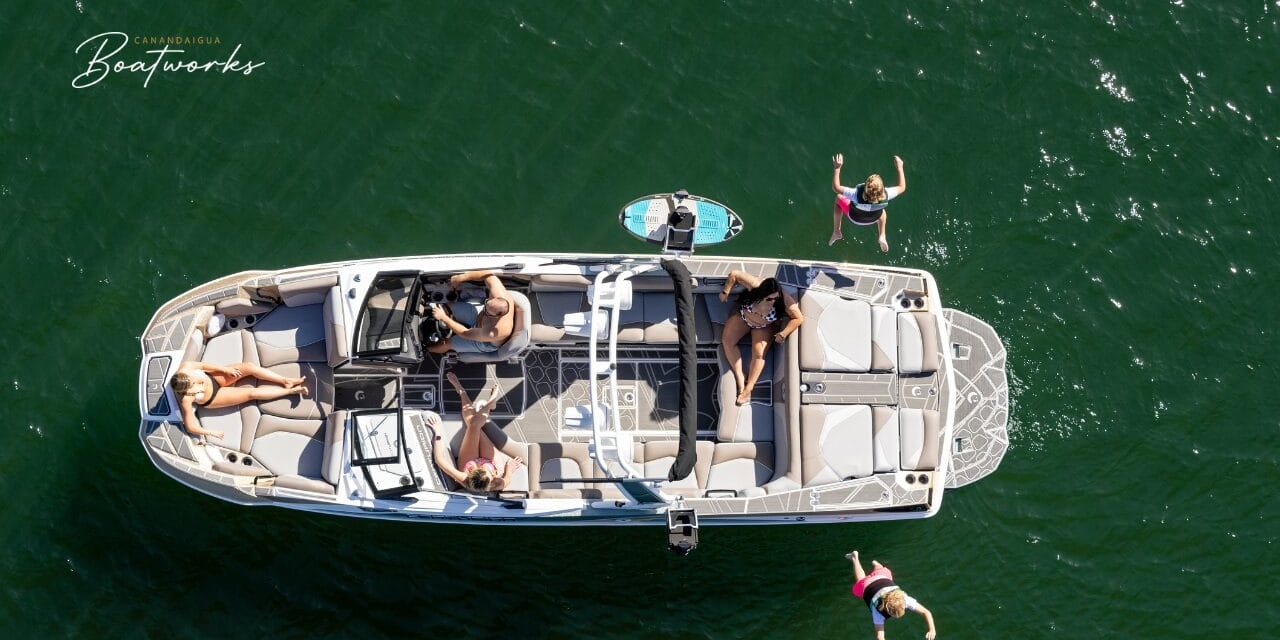 Aerial view of a boat on the water with four people. Two are seated inside, one is standing at the rear, and one person is jumping into the water.
