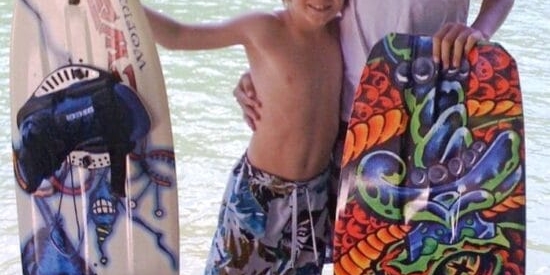 Two kids are standing on a dock by a lake, each holding a wakeboard. The younger child is shirtless, and the older child is wearing a white shirt. Trees are visible across the water.