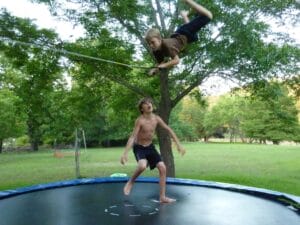 Two boys are on a trampoline. One is mid-air holding a rope swing. The other is standing on the trampoline. Trees and grass are in the background.