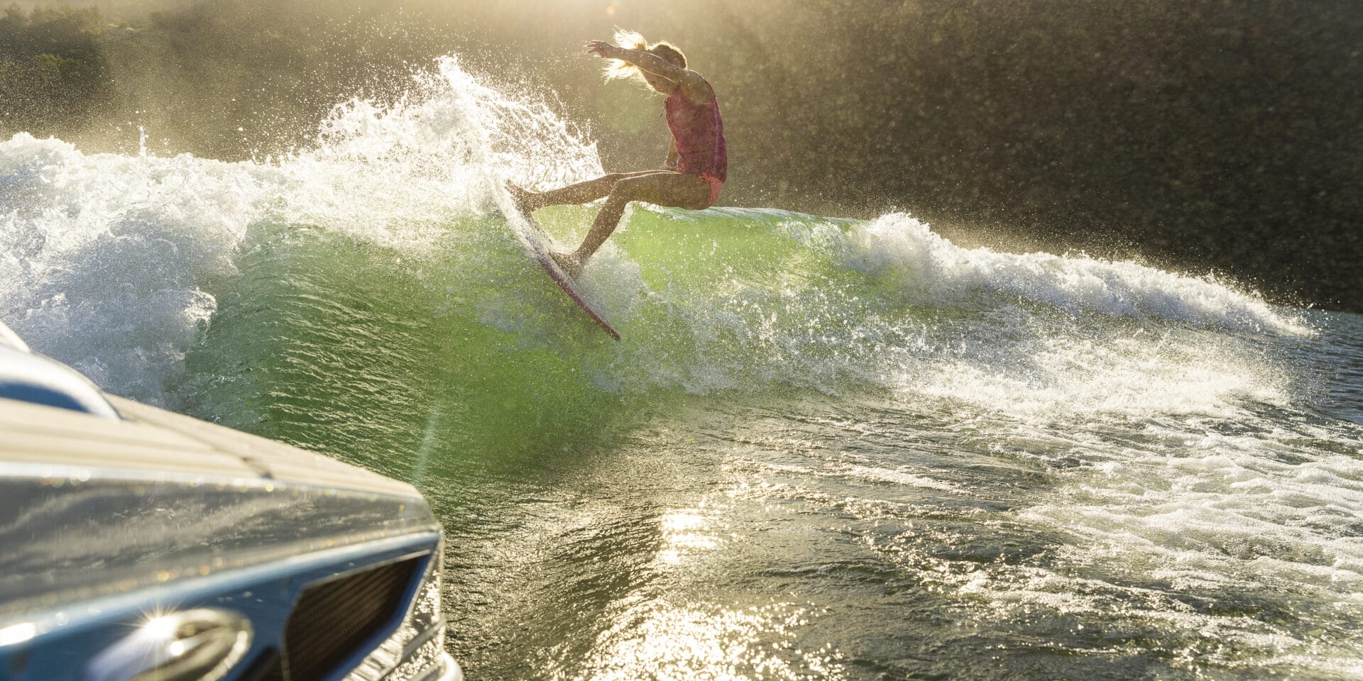 A person surfs on a wave behind a boat in a sunny outdoor setting with a forested hillside in the background.