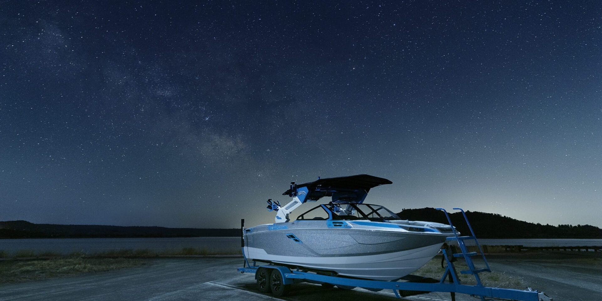 A boat on a trailer is parked on a lakeside road at night, with a starry sky in the background.