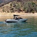 A motorboat with a black canopy cruises on a calm lake, with a tree-covered hillside in the background on a sunny day.