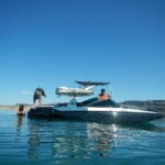 A person dives into the water from a boat, while another person sits at the helm. A third person is in the water near the boat on a sunny day.