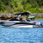 A sleek motorboat on a lake, featuring a canopy and a wakeboard attached at the back, with trees and a wooden pier in the background.