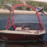 Red and white boat with a wakeboard tower floats on a calm lake near the shore. Two wakeboards and speakers are attached to the tower.