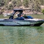 A sleek motorboat with a metallic finish is navigating a greenish lake, with rocky and tree-lined shorelines in the background.