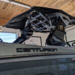 A Centurion boat is inside a garage, featuring a shiny exterior and a black metal structure above. The ceiling has wooden beams and there are fluorescent lights.