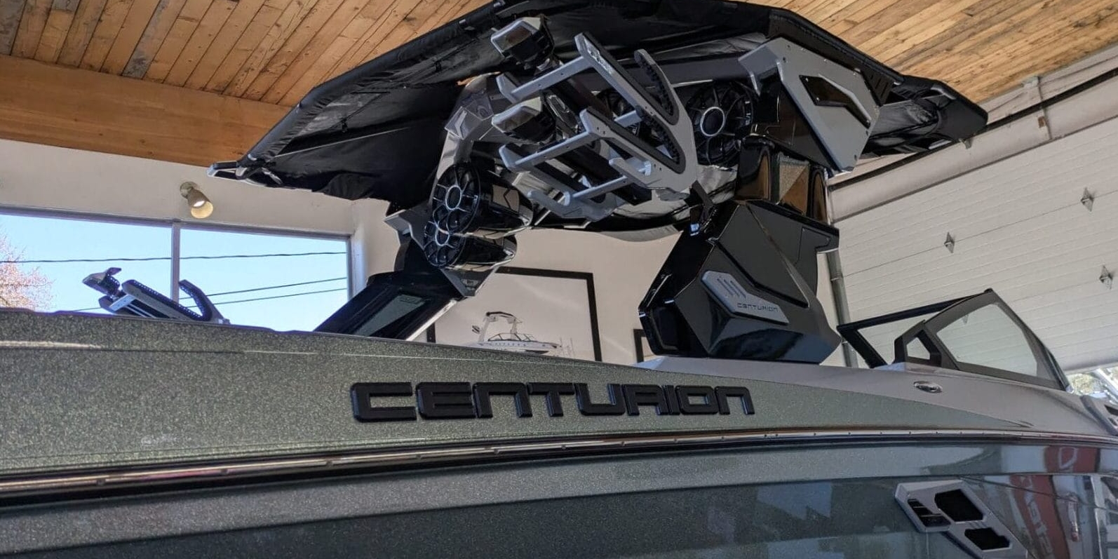 A Centurion boat is inside a garage, featuring a shiny exterior and a black metal structure above. The ceiling has wooden beams and there are fluorescent lights.