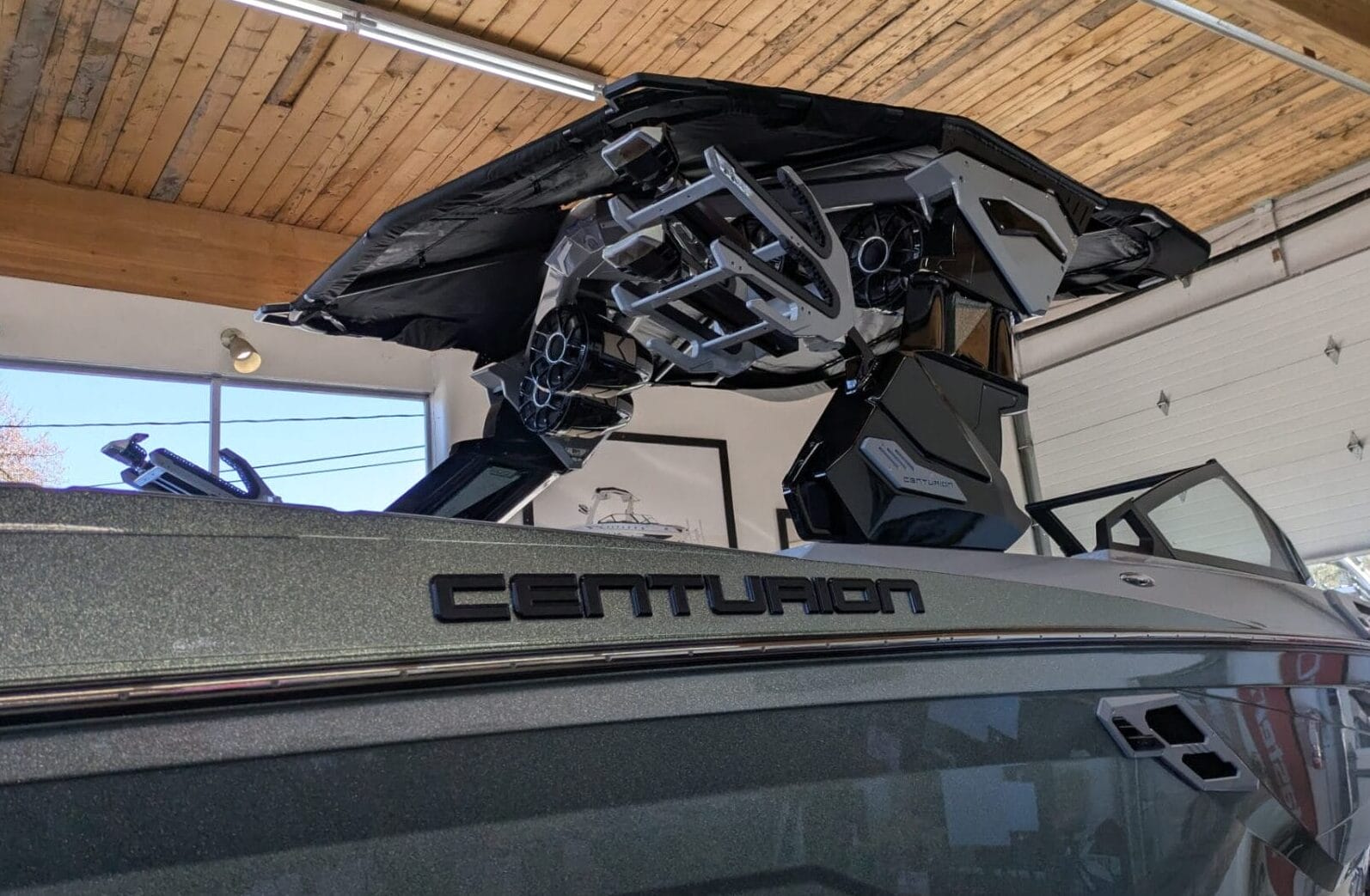 A Centurion boat is inside a garage, featuring a shiny exterior and a black metal structure above. The ceiling has wooden beams and there are fluorescent lights.