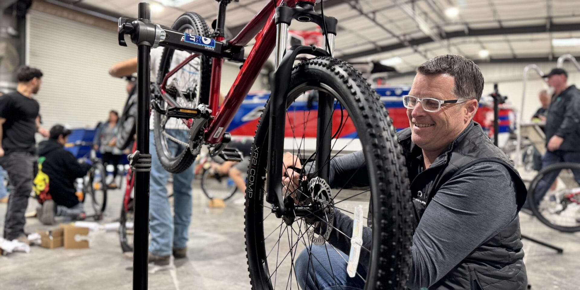 Man in a workshop adjusting the front wheel of a red mountain bike on a repair stand. Other people and bicycles are in the background.