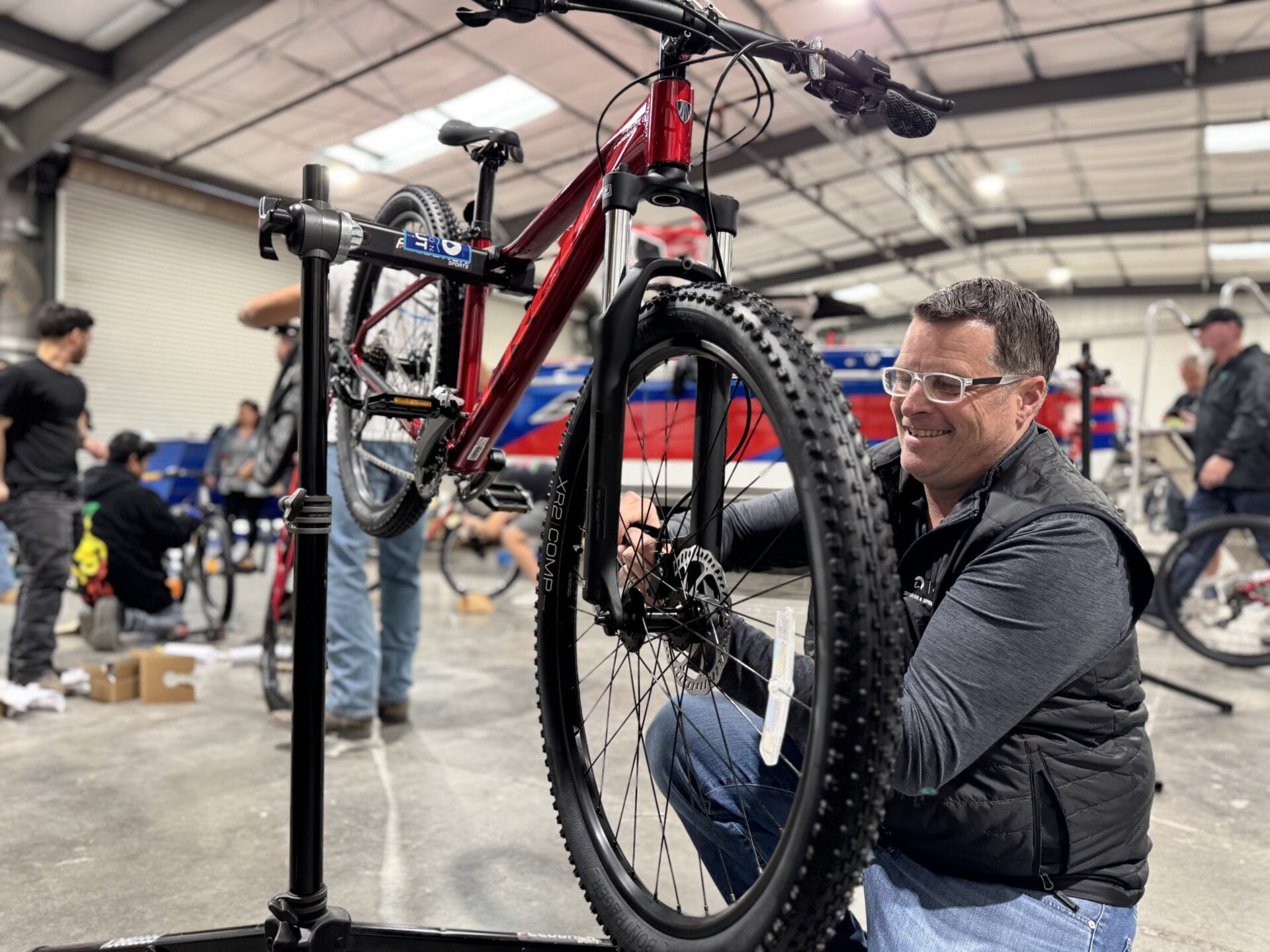Man in a workshop adjusting the front wheel of a red mountain bike on a repair stand. Other people and bicycles are in the background.