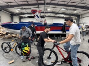 Four men are working on bicycles in a spacious garage with a large blue, white, and red boat in the background.