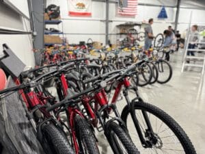 Several rows of red bicycles are lined up in a warehouse. People are in the background, some working on assembling bikes. American and California flags hang on the wall.