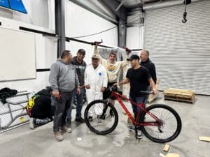 Seven people in a warehouse gather around a red bicycle. Two wear lab coats, and one holds the bike. Piles of materials are visible in the background.