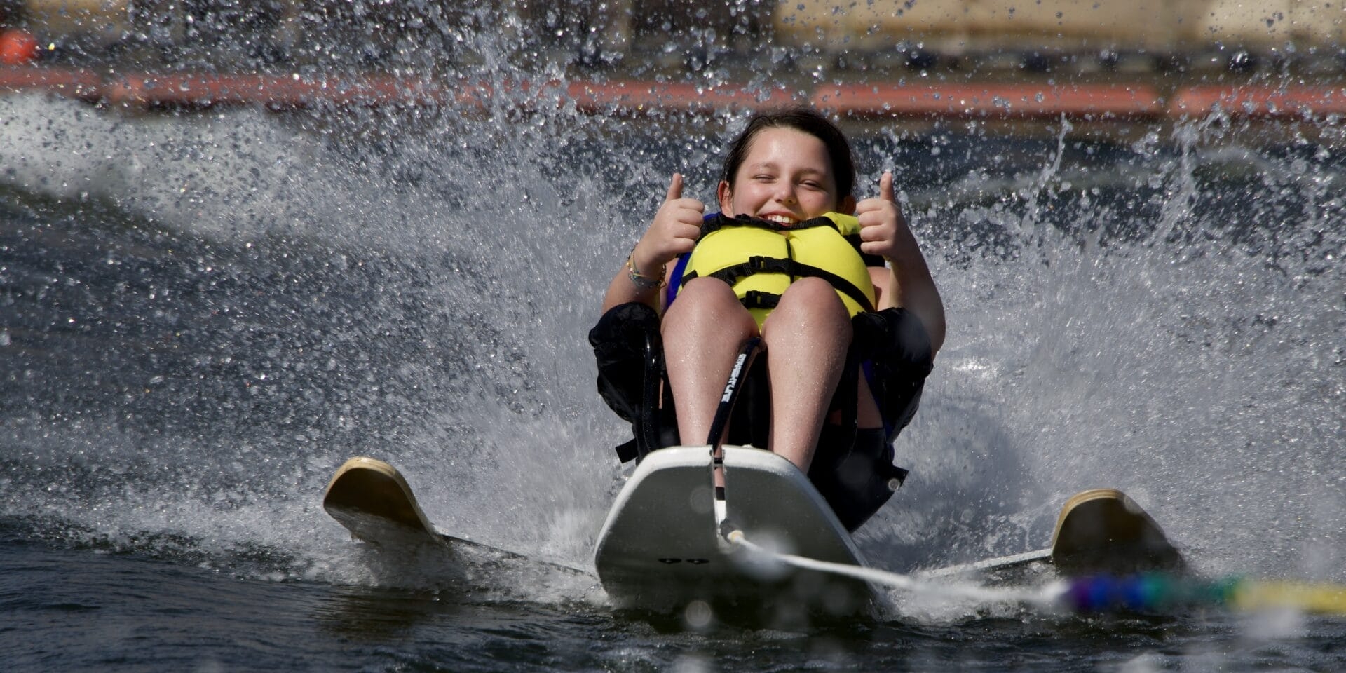 A person enjoys water skiing, sitting on a ski board, wearing a life jacket, and giving a thumbs-up with both hands as water splashes around.