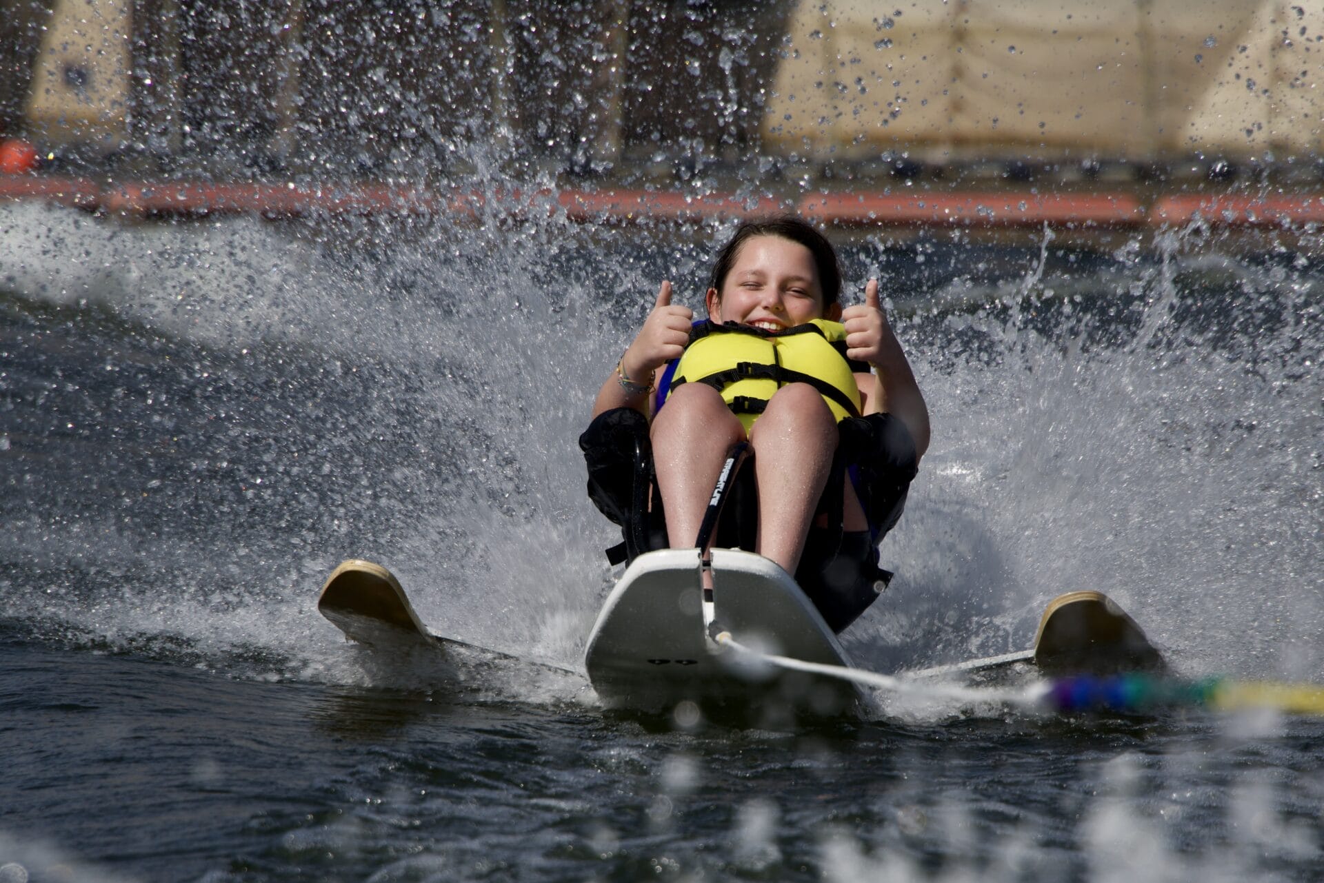 A person enjoys water skiing, sitting on a ski board, wearing a life jacket, and giving a thumbs-up with both hands as water splashes around.