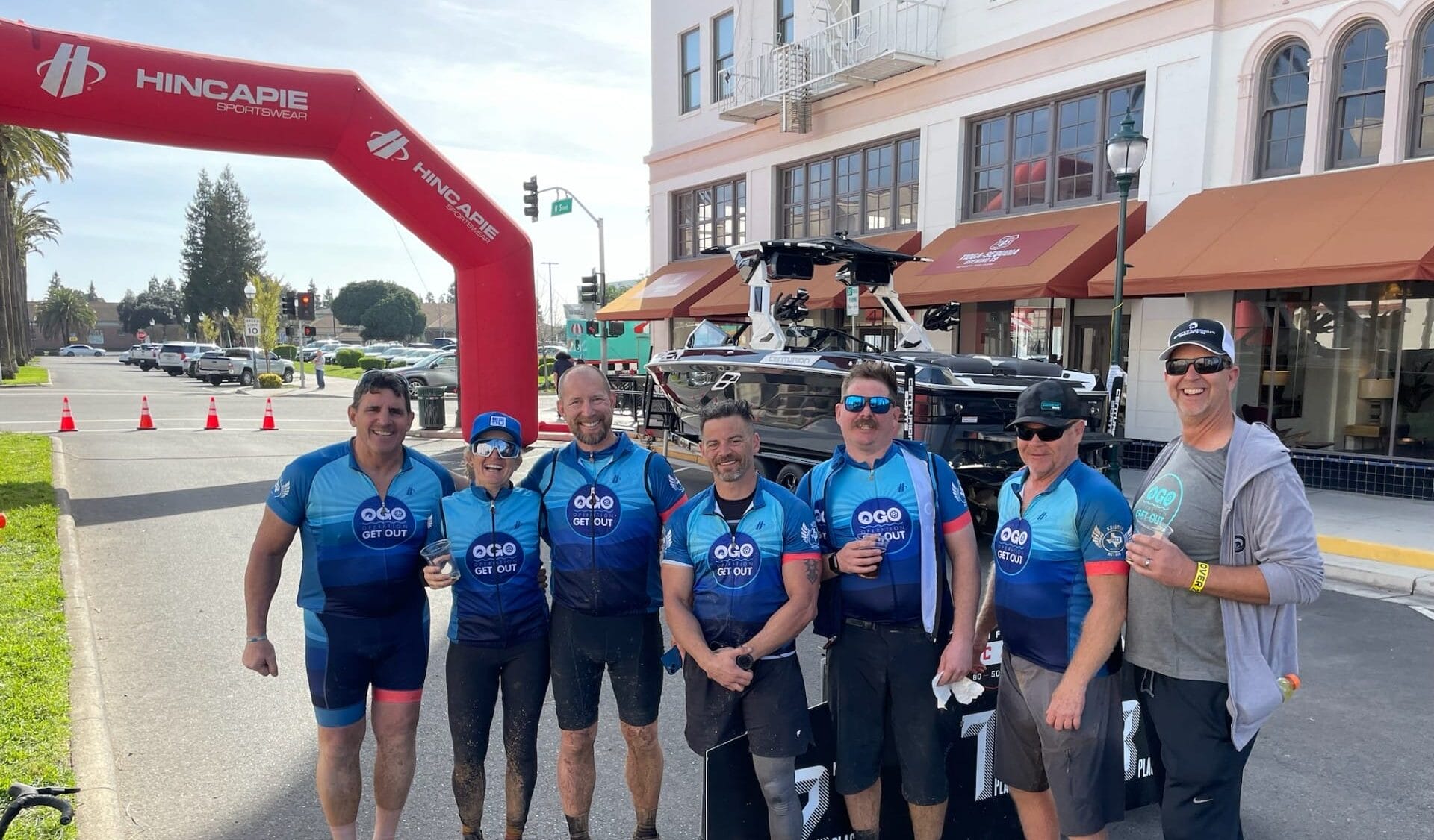 Seven cyclists in matching blue jerseys pose together under an arch with bicycles in the background. Some are barefoot. They're smiling and holding drinks.
