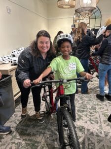 Woman and child with bicycle and helmet, smiling at camera. People and bicycles in background.
