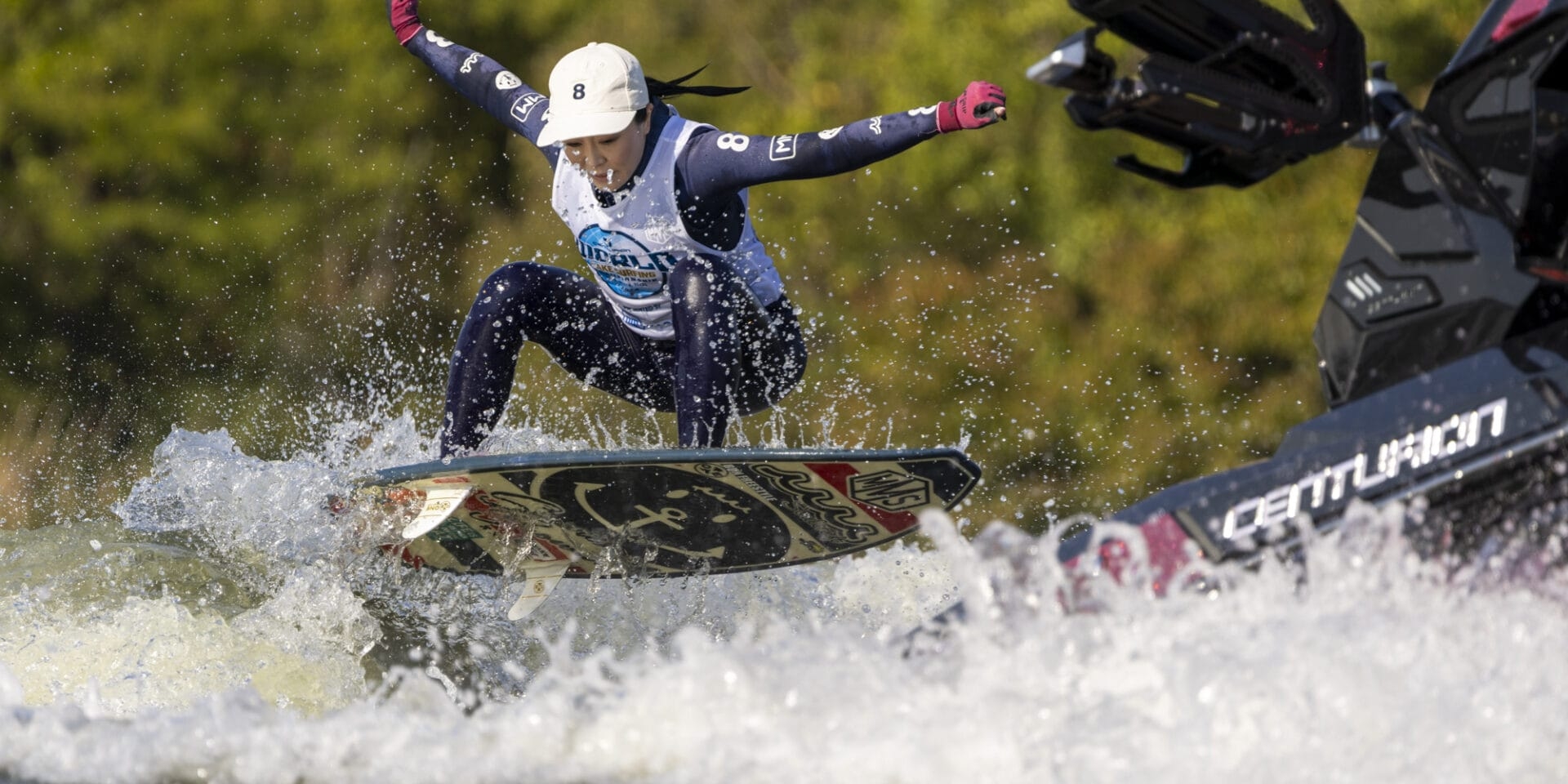 A person wake surfing on a lake with a wakeboard, wearing a life vest and helmet, while a boat creates waves nearby.