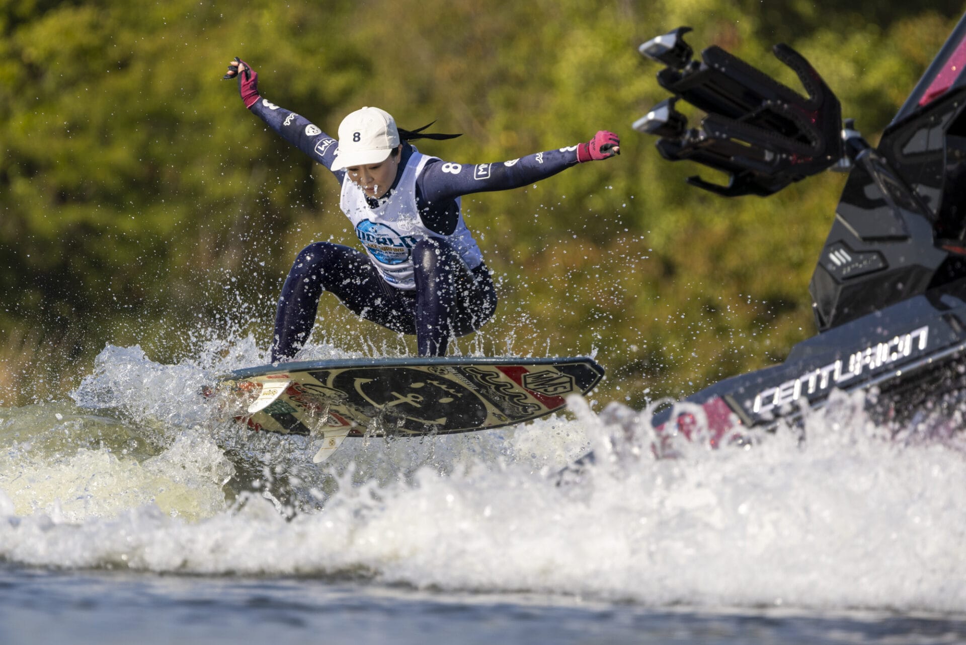 A person wake surfing on a lake with a wakeboard, wearing a life vest and helmet, while a boat creates waves nearby.