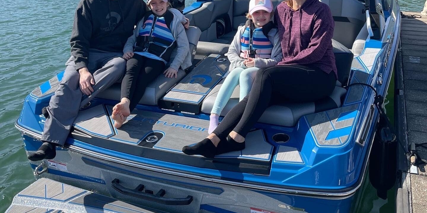 Family of four sitting on a blue boat docked at a lakeside, all wearing casual clothes and sunglasses. Two children in life jackets. Trees and houses visible in the background.