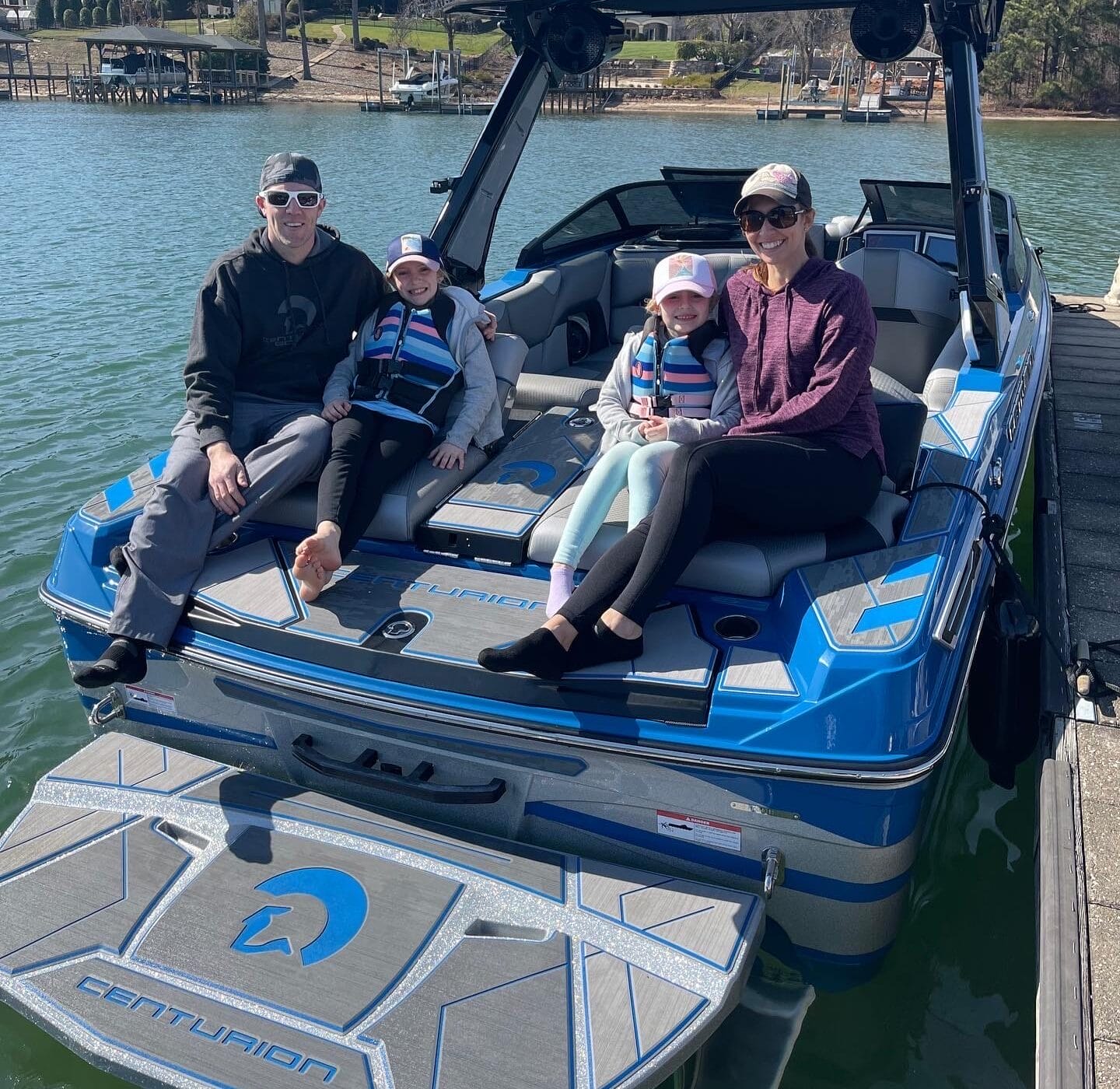 Family of four sitting on a blue boat docked at a lakeside, all wearing casual clothes and sunglasses. Two children in life jackets. Trees and houses visible in the background.