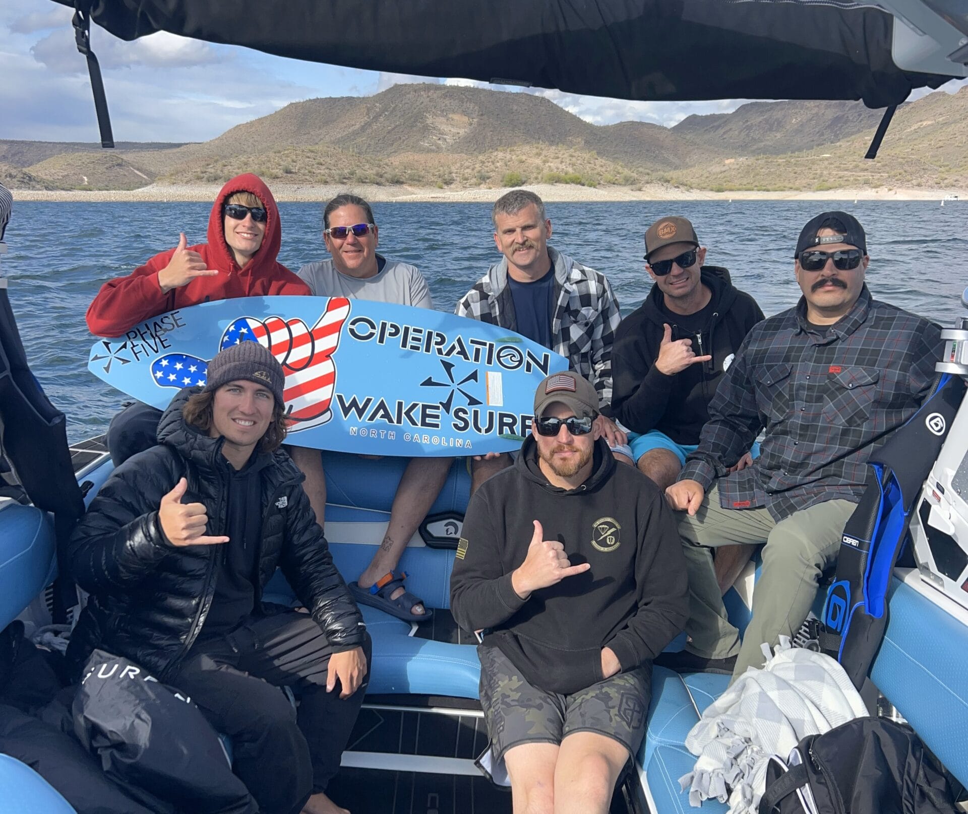 Group of seven men on a boat holding a "Operation Wake Surf" surfboard, with hills and water in the background. Some make hand gestures, wearing casual and sporty attire.
