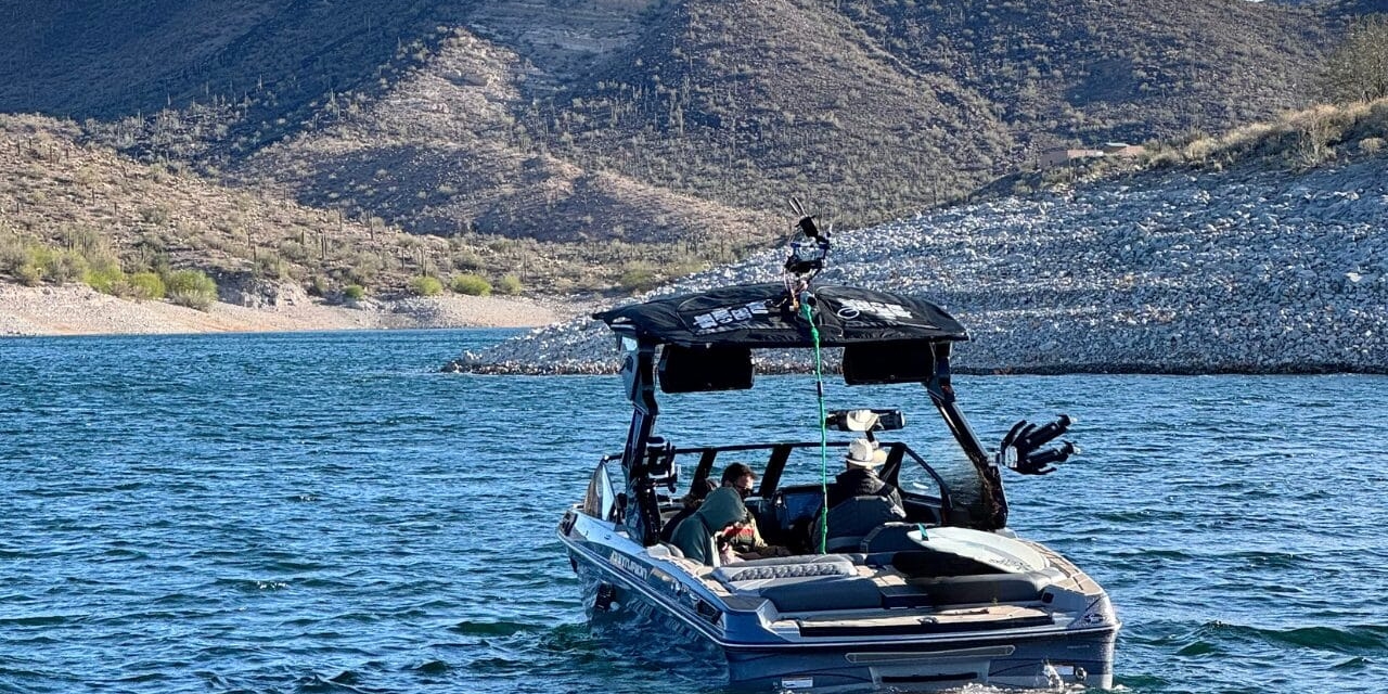 A motorboat with a canopy is on a lake, with a rocky shore and hills in the background. Three people are onboard.
