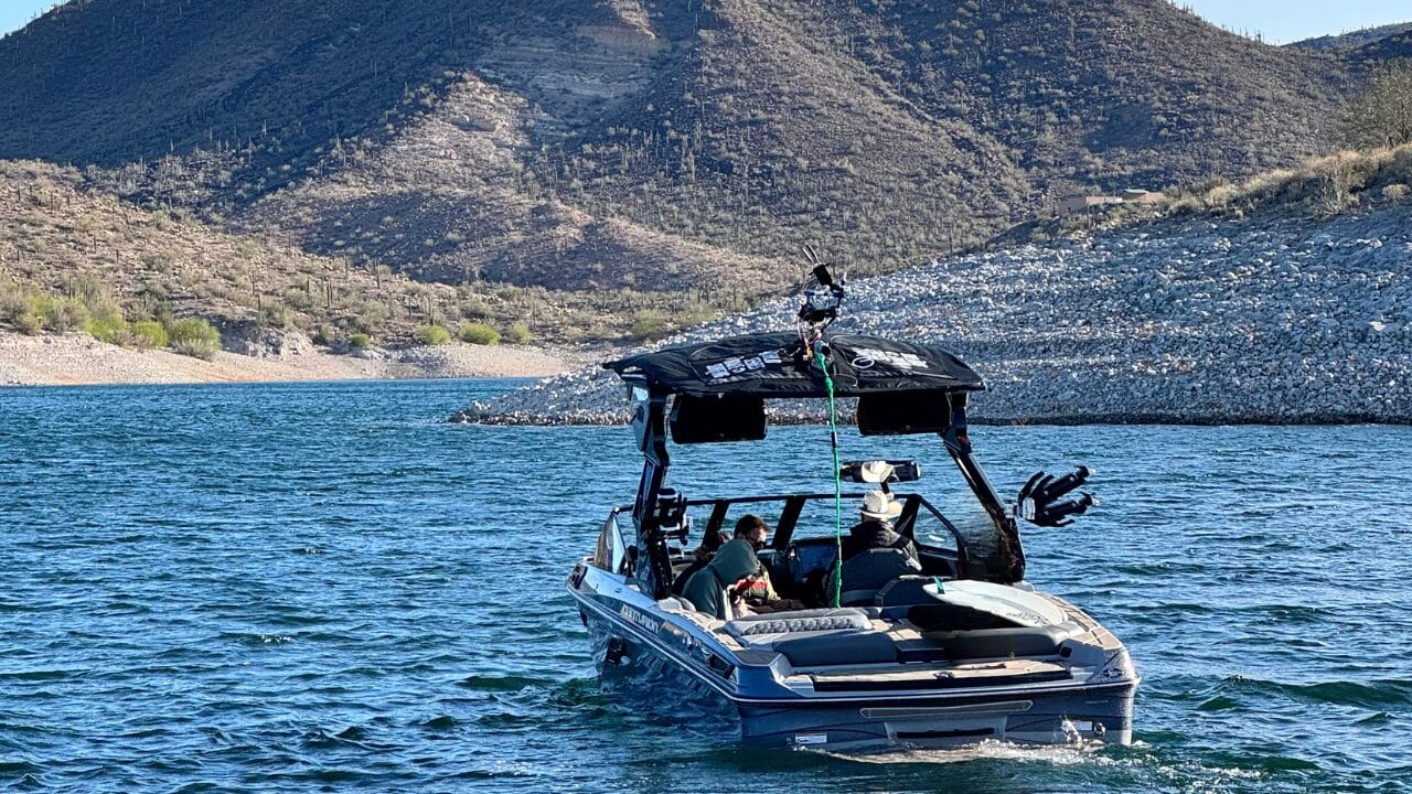 A motorboat with a canopy is on a lake, with a rocky shore and hills in the background. Three people are onboard.
