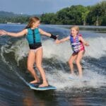 Two young girls wearing life vests high-five while surfing beside each other on a river, with trees and a distant shoreline in the background.