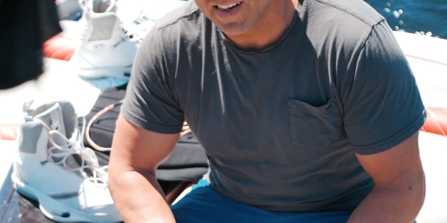 A man wearing a camouflage hat, sunglasses, grey t-shirt, and blue shorts sits on a boat near water sports gear with water in the background.