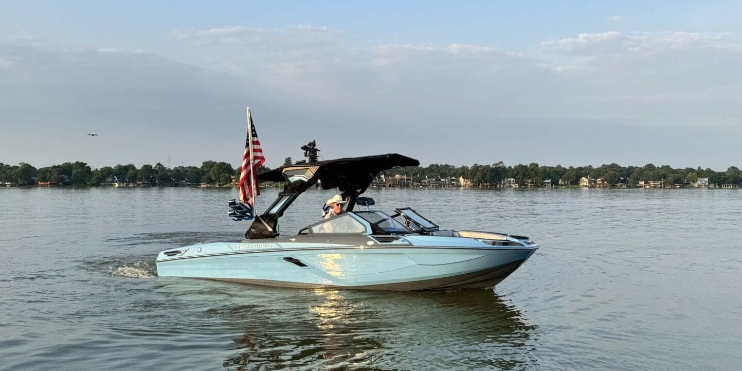 A light blue motorboat with an American flag moves across a calm lake under a partly cloudy sky, with a tree-lined shore in the background.